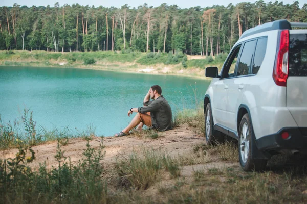 Homem sentado perto de carro branco SUV na borda olhando para o lago com água azul — Fotografia de Stock