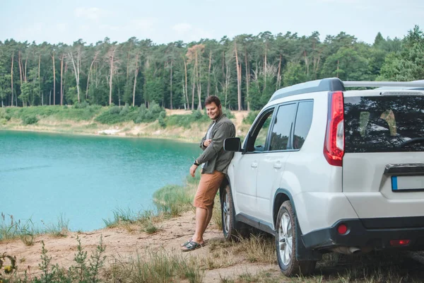 Homem perto de carro branco SUV na borda olhando para o lago com água azul — Fotografia de Stock