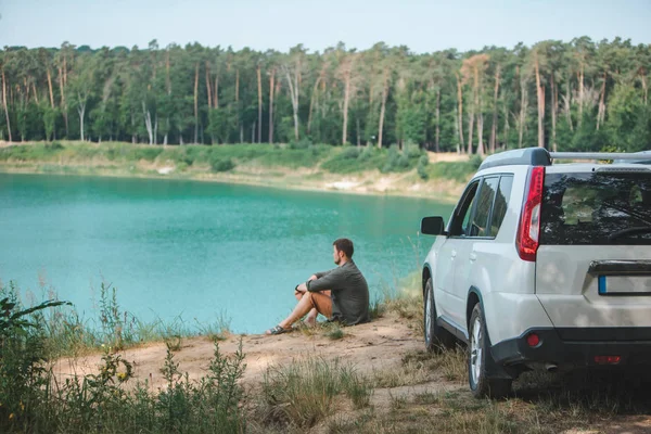 Homem sentado perto de carro branco SUV na borda olhando para o lago com água azul — Fotografia de Stock