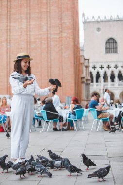Beyaz giysili, hasır şapkalı kadın Venedik Meydanı 'nda güvercinlerle eğleniyor.
