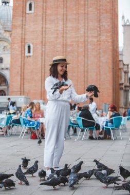 Beyaz giysili, hasır şapkalı kadın Venedik Meydanı 'nda güvercinlerle eğleniyor.