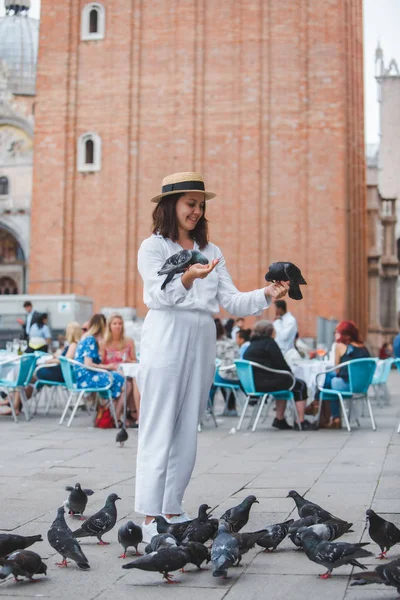 Beyaz giysili, hasır şapkalı kadın Venedik Meydanı 'nda güvercinlerle eğleniyor.