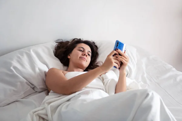 Жінка зі смартфоном у ліжку з білими простирадлами — стокове фото