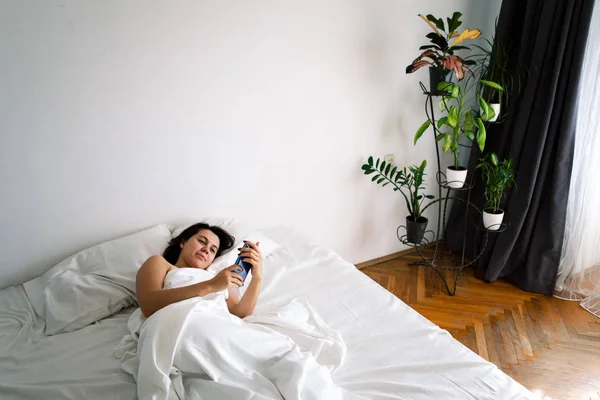 Mulher com smartphone na cama com lençóis brancos — Fotografia de Stock