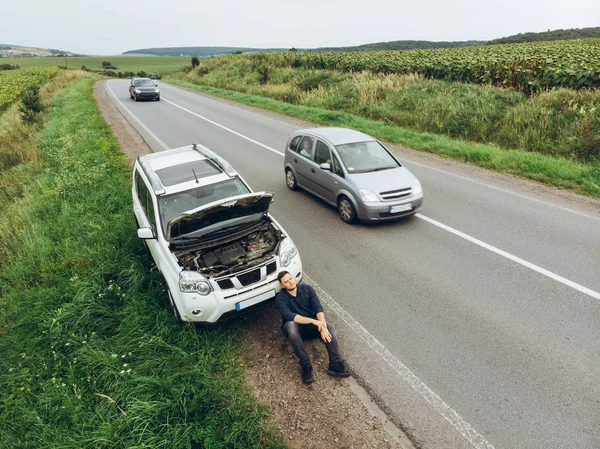 Hombre sentado en la carretera cerca de coche roto. tratando de detener el coche de ayuda —  Fotos de Stock