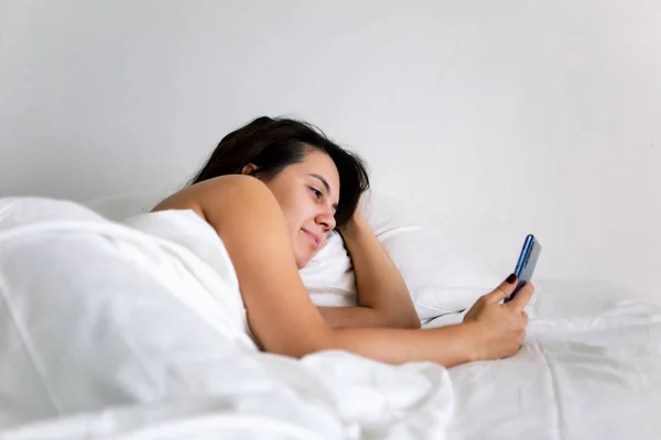 Woman with smartphone in bed with white sheets — Stock Photo, Image