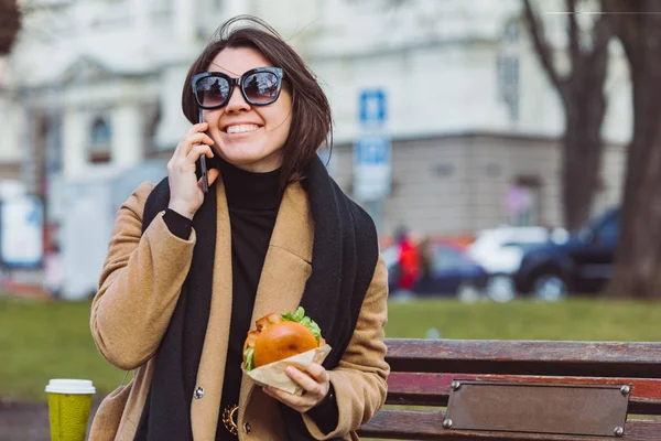 Young beauty business woman eating fast food and working on phone while sitting at city bench — Stock Photo, Image