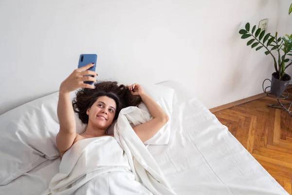 Woman laying in bed taking selfie on the phone — Stock Photo, Image