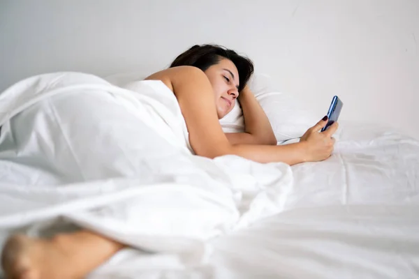 Жінка зі смартфоном у ліжку з білими простирадлами — стокове фото