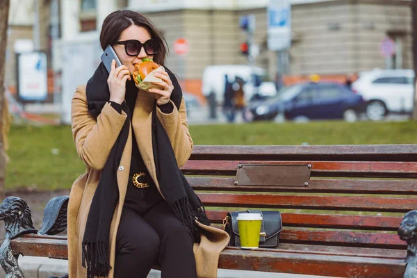 Young beauty business woman eating fast food and working on phone while sitting at city bench — Stock Photo, Image