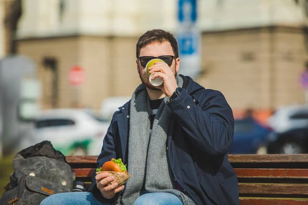 Jovem homem sorrindo comer hambúrguer e beber café sentado no banco da cidade — Fotografia de Stock