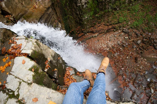Женщина в синих джинсах и коричневых сапогах сидит на краю, глядя на водопад — стоковое фото