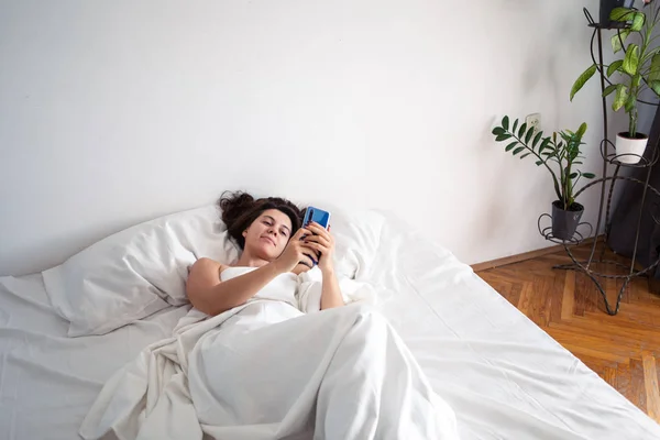 Женщина со смартфоном в постели с белыми простынями — стоковое фото