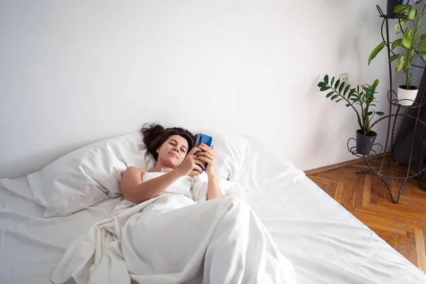 Woman with smartphone in bed with white sheets — Stock Photo, Image