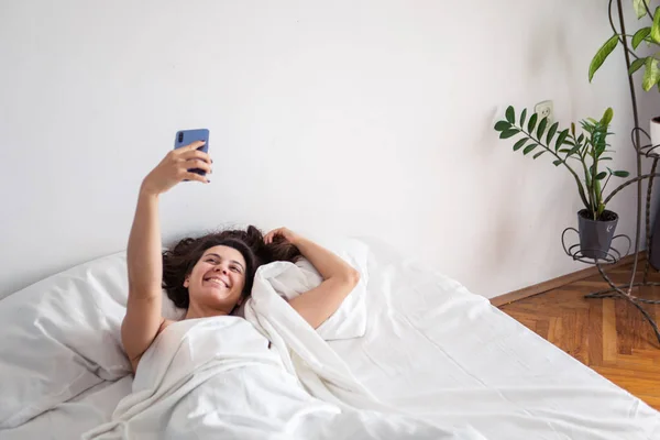 Женщина, лежащая в постели и делающая селфи по телефону — стоковое фото