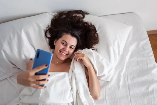 Женщина, лежащая в постели и делающая селфи по телефону — стоковое фото