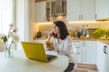 mutfakta oturan kadın telefonda çay içerken konuşurken dizüstü bilgisayar üzerinde çalışıyor