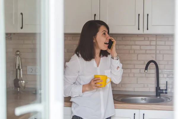 Smiling happy woman talking no phone drinking tea from yellow mug at the kitchen — Stock Photo, Image