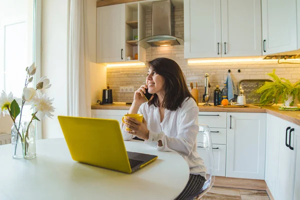 Mulher sentada na cozinha trabalhando no laptop falando sobre o telefone beber chá — Fotografia de Stock
