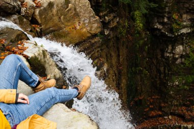 mavi kot pantolon ve kahverengi çizmeler şelale bakarak kenarında oturan kadın