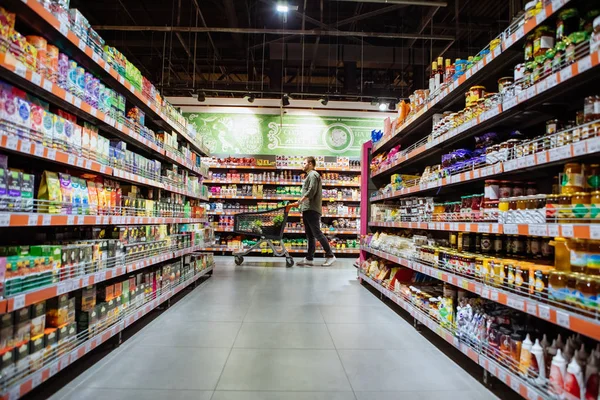 Jovem com carrinho de compras entre prateleira da loja — Fotografia de Stock