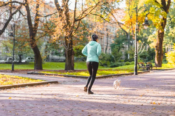 女人在秋天的城市公园用皮带与狗赛跑 — 图库照片