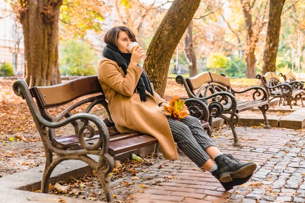 Woman sitting on the bench at autumn city park drinking coffee — Stock Photo, Image