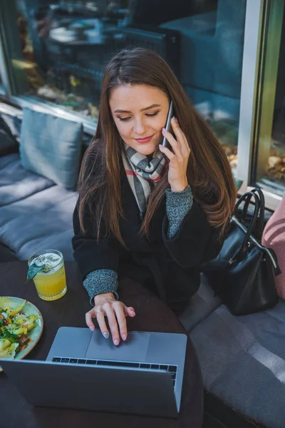 Kafede dizüstü bilgisayar üzerinde çalışan genç güzel iş kadını telefonda konuşurken — Stok fotoğraf