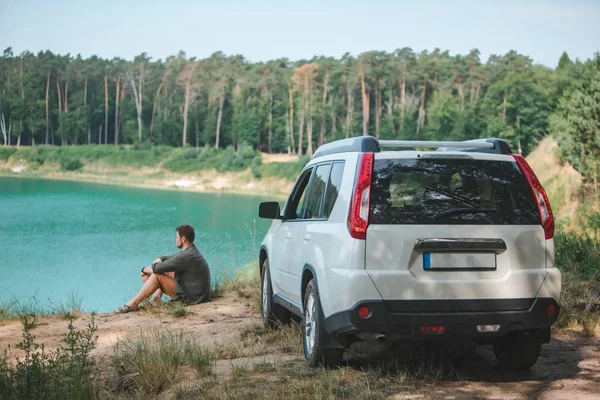 Hombre sentado cerca de coche suv blanco en el borde mirando al lago con agua azul — Foto de Stock