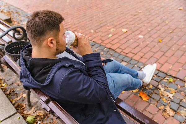 Homem sentado no banco no parque da cidade beber café surf no telefone — Fotografia de Stock