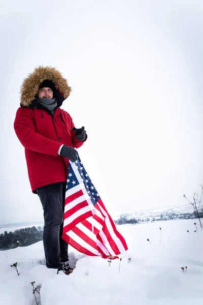 Ο άνθρωπος στο κόκκινο χειμερινό παλτό κρατώντας την σημαία των ΗΠΑ σε εξωτερικούς χώρους σε χιονισμένο καιρικές συνθήκες — Φωτογραφία Αρχείου