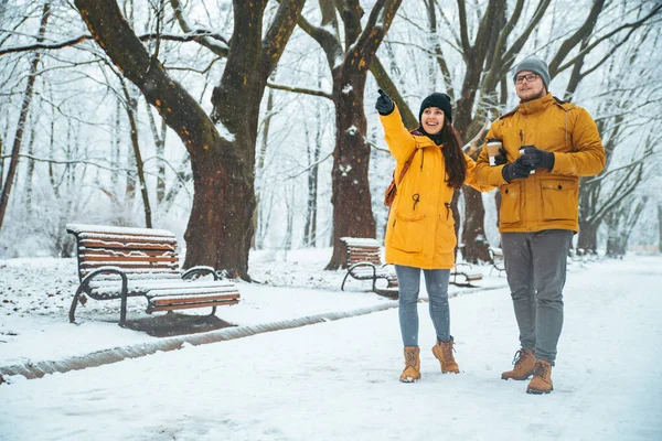 Pareja caminando por el parque de la ciudad nevada bebiendo café para ir señalando algo recto — Foto de Stock