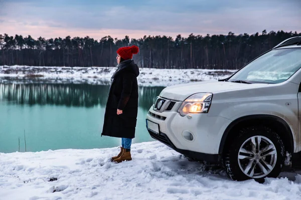 Mladá dospělá žena v zimním oblečení s červeným kloboukem s bubo stojící v blízkosti SUV auto na řece straně s krásným výhledem — Stock fotografie