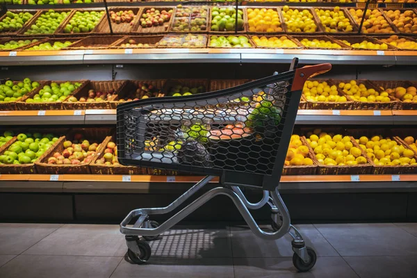 Shopping cart in the middle of the picture shelf with oranges and apples on background — Stock Photo, Image