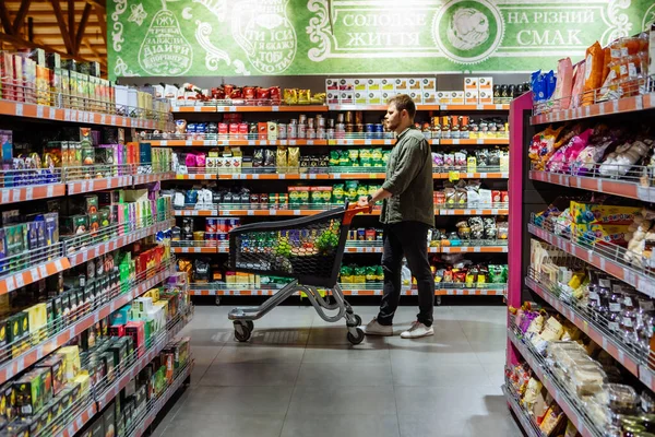 Jovem com carrinho de compras entre prateleira da loja — Fotografia de Stock