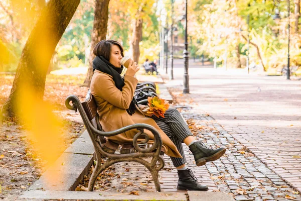 Woman sitting on the bench at autumn city park drinking coffee — Stock Photo, Image