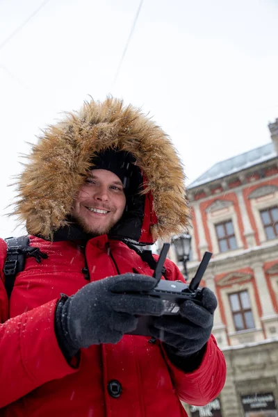 Retrato de hombre adulto joven en la capucha con piel en la plaza de la ciudad de invierno con controlador de drones en las manos — Foto de Stock