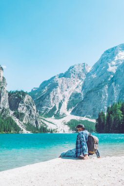 Arka planda sırt çantalı dağlarla gölün sahilinde oturan adam dolomitler.