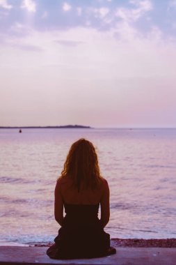 Kadın silueti sahilde oturmuş denizin üzerinde gün batımını seyrediyor. Boşluğu kopyala