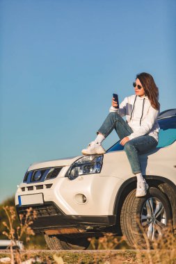 Beyaz arazi aracının yanındaki kadın günbatımı yolculuğunun tadını çıkarıyor. Arka planda mavi gökyüzü.