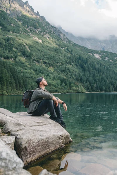 山の中の湖の前の岩の上に座ってハイキングのコンセプトを楽しんでいる男 — ストック写真