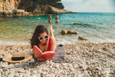 Kırmızı mayo giyen kadın deniz sahilinde battaniyeye uzanmış güneşlenirken kitap okuyor.
