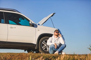 Gün batımında kaputu açık bir şekilde arabanın yanında oturan bir adam telefonla fotokopi uzayı hakkında konuşuyor.