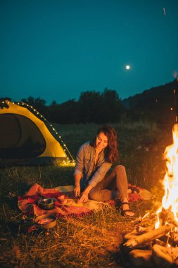 Genç ve güzel bir kadın ateşin önünde yemek pişiriyor. Arka planda sarı çadır. Boşluğu kopyala