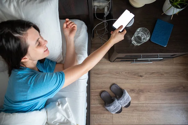 Awake Woman Taking Phone Wireless Charger Bedside Table Overhead Top — Stock Photo, Image