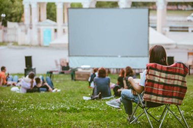 Kamp koltuğunda oturan kadın açık hava sinemasında film izliyor. Patlamış mısır yiyip gazoz içiyorum.