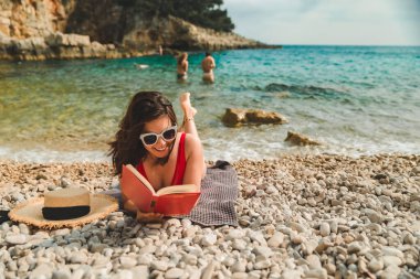 Kırmızı mayo giyen kadın deniz sahilinde battaniyeye uzanmış güneşlenirken kitap okuyor.