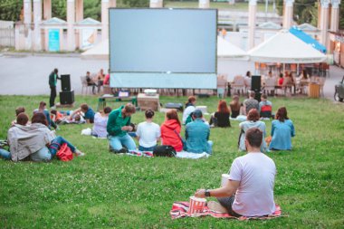 Şehir parkındaki açık hava sinemasında battaniyenin üzerinde oturan adam büyük ekranın önünde abur cubur yiyor.