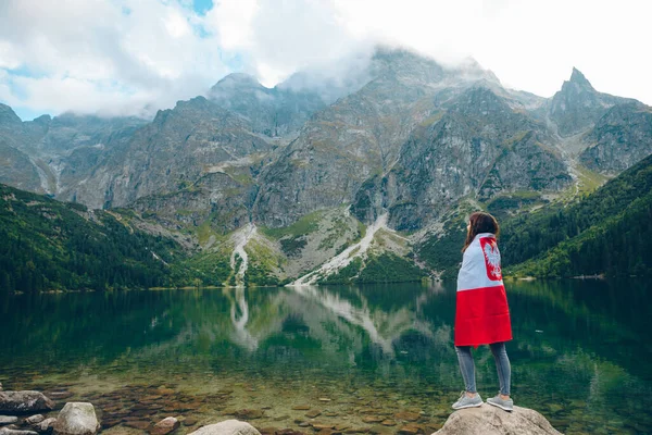 Mujer joven cubierta con bandera de Polonia mirando al lago en las montañas de Tatra — Foto de Stock