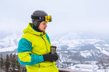 Adam kar kayağı kıyafeti kazandı. Doldurulabilir bardaktan sıcak çay içerek arka plandaki kar dağlarına baktı.